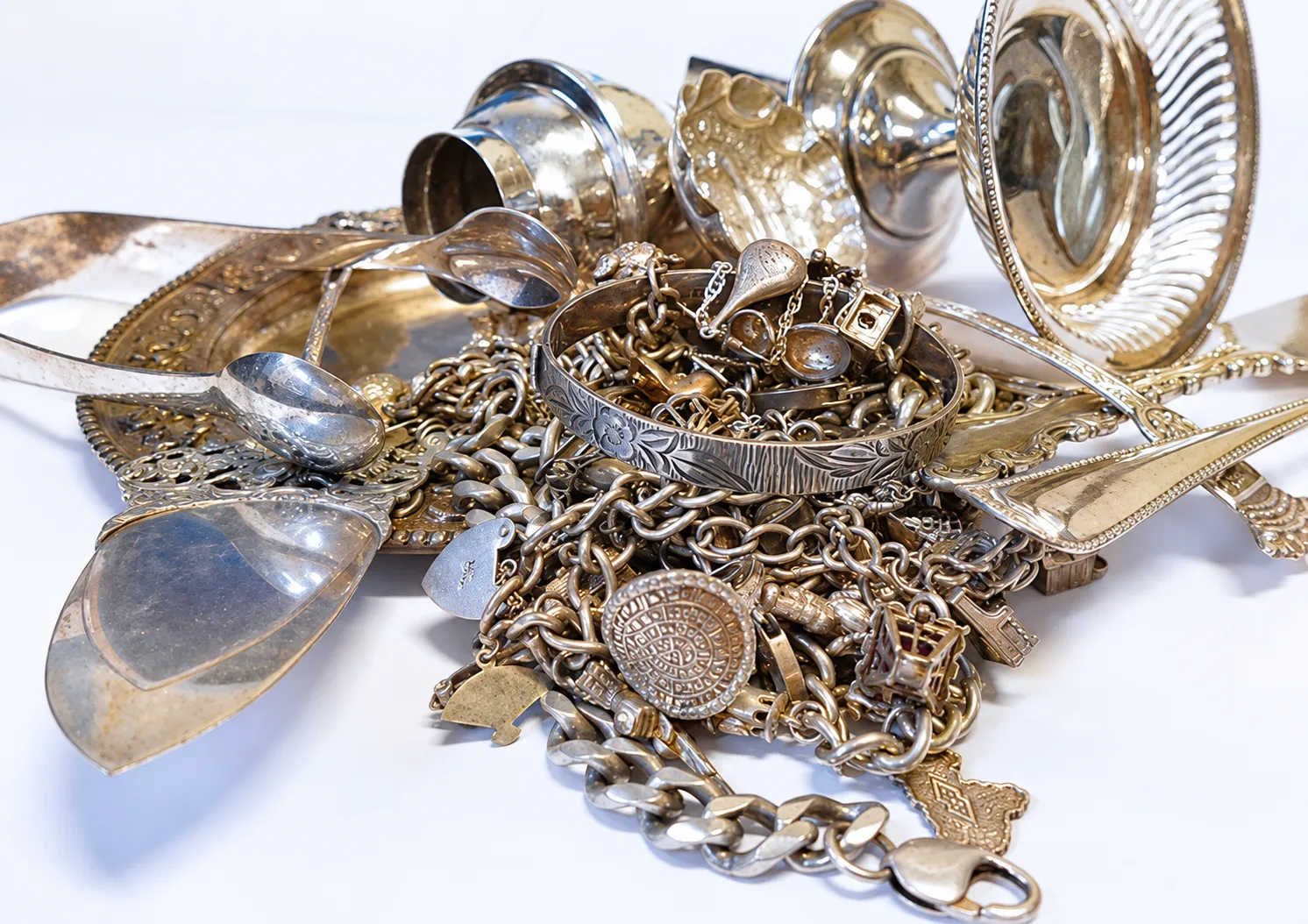 Montón de piezas de plata sobre fondo blanco: cucharas y cubiertos, una pulsera grabada, cadenas gruesas, dijes y pequeñas copas y bandejas.